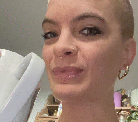 Patientin mit kurzen, rasierten Haaren hält eine Tasse und lächelt leicht in die Kamera, im Hintergrund eine gemütliche Wohnungseinrichtung