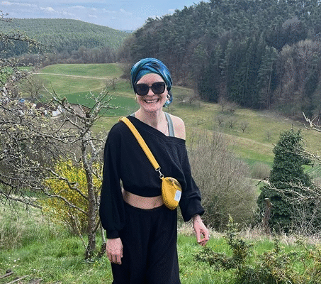 Patientin mit Kopftuch, Sonnenbrille und sportlicher Kleidung steht lachend bei einer Wanderung in hügeliger Landschaft mit Wald und Wiesen