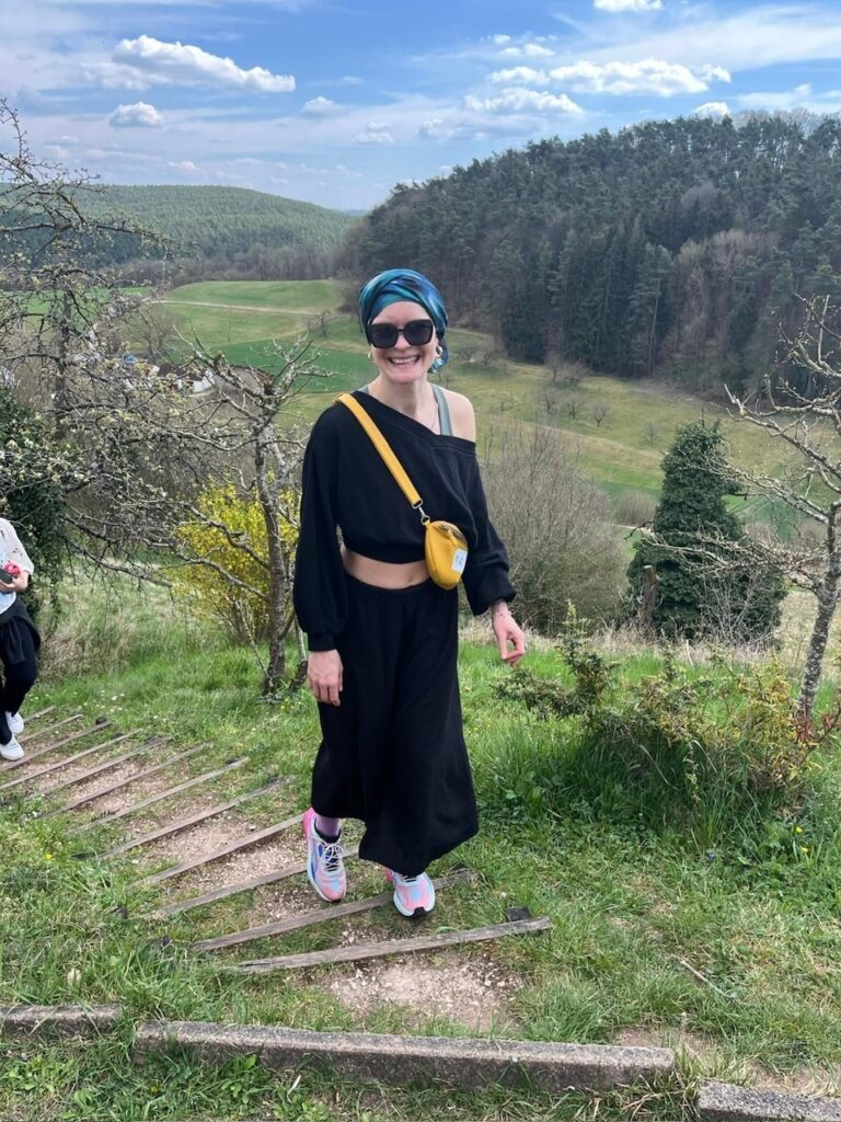Patientin mit Kopftuch, Sonnenbrille steht lachend bei einer Wanderung in hügeliger Landschaft mit Wald und Wiesen