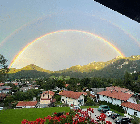 Doppelter Regenbogen spannt sich über ein Dorf mit roten Dächern und umliegenden grünen Bergen, im Vordergrund rote Blumen