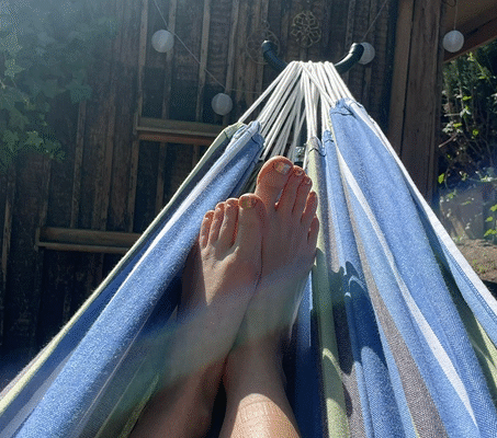 Füße einer Patientin in einer blauen Hängematte, Sonnenstrahlen fallen durch die Blätter, im Hintergrund eine Holzwand und Gartenatmosphäre